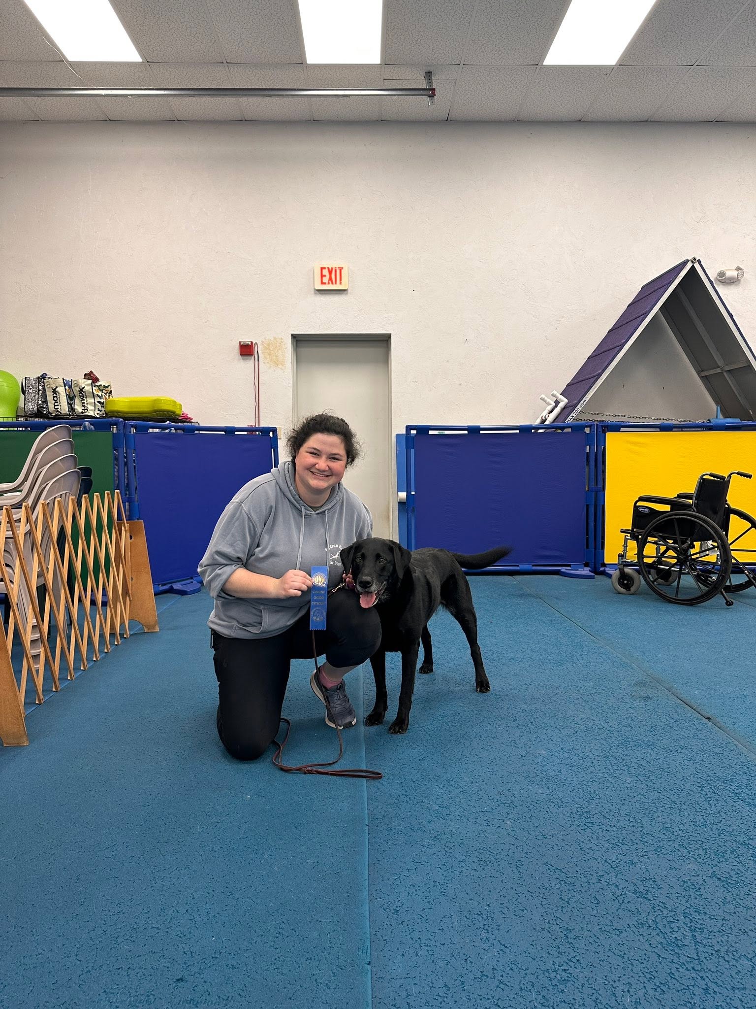 Emily kneeling beside a black Labrador indoors with a blue ribbon