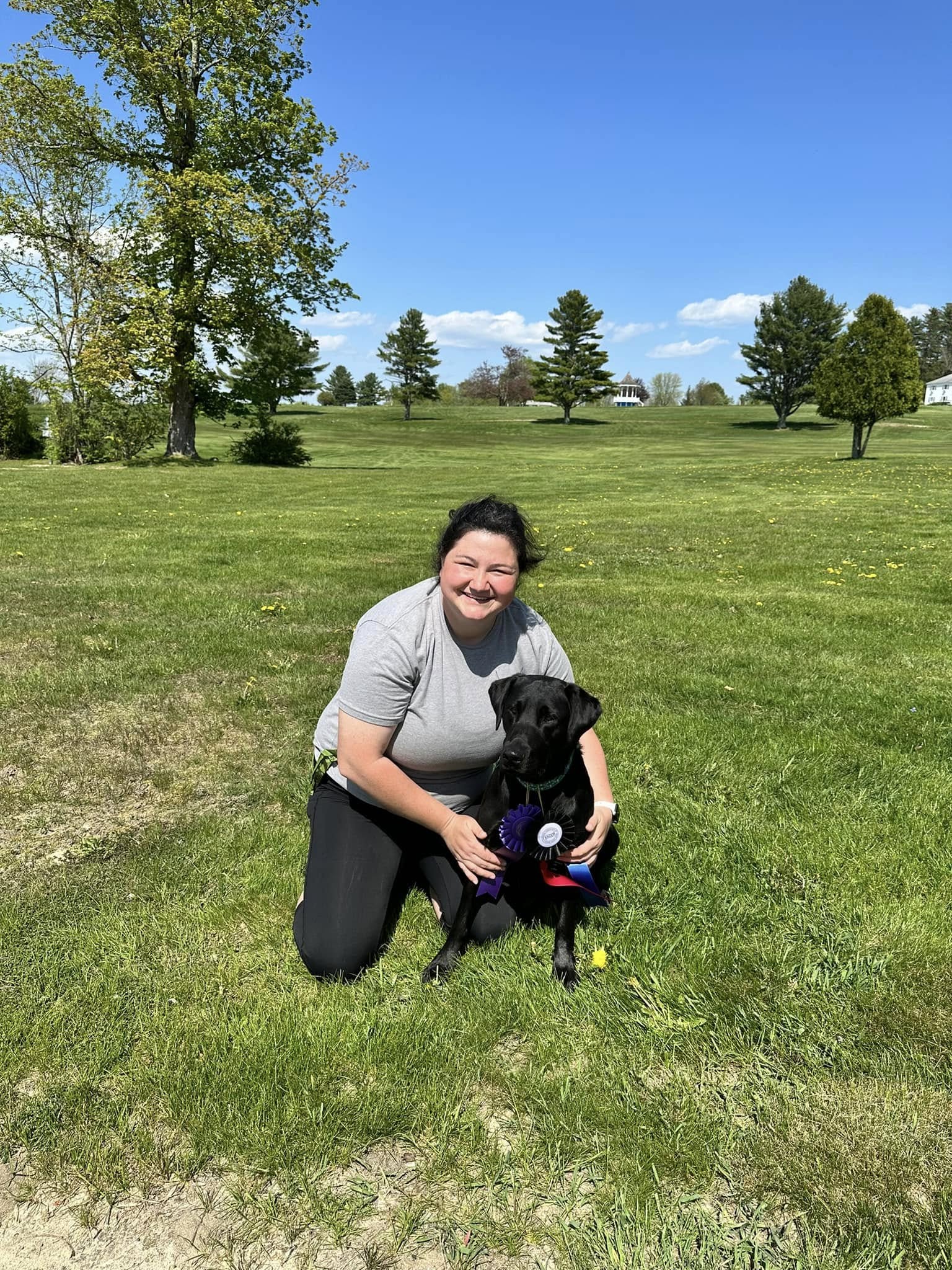 Emily kneeling with a black Labrador wearing ribbons on a grassy field