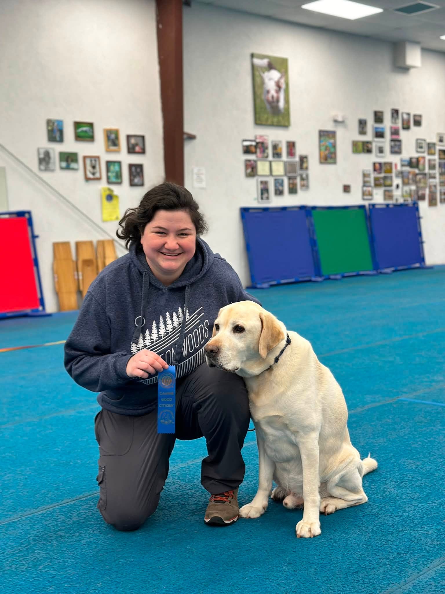 Emily kneeling beside a yellow Labrador and holding a blue ribbon indoors
